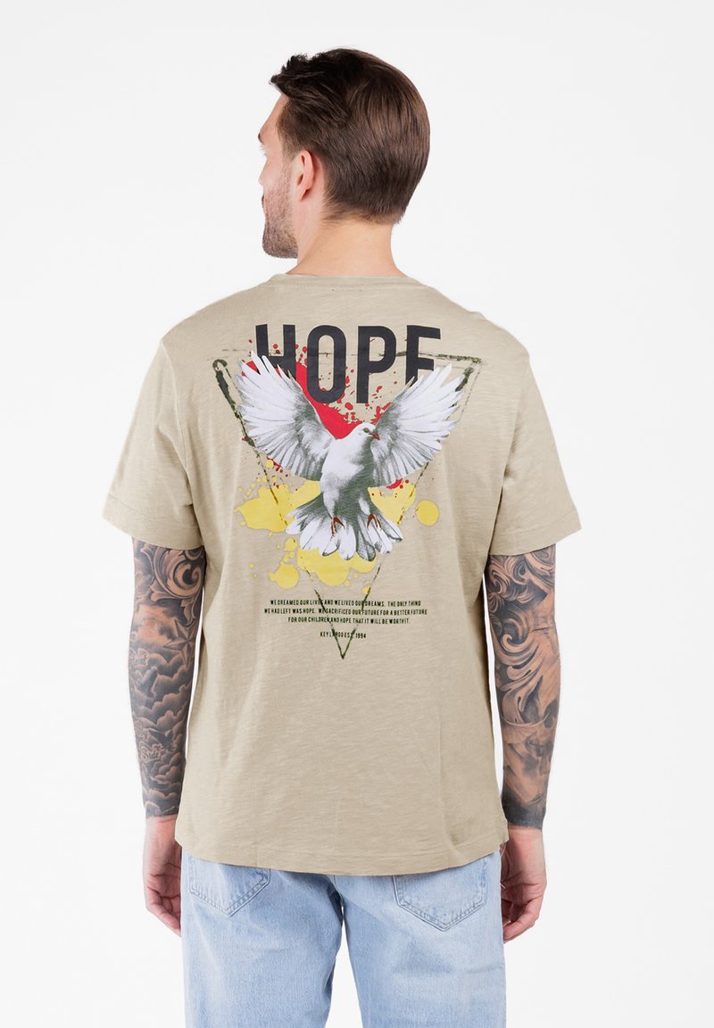 Hombre con los brazos tatuados lleva una camiseta beige con una paloma blanca, manchas de pintura y el texto "ESPERANZA" en la espalda, combinada con jeans de color azul claro.