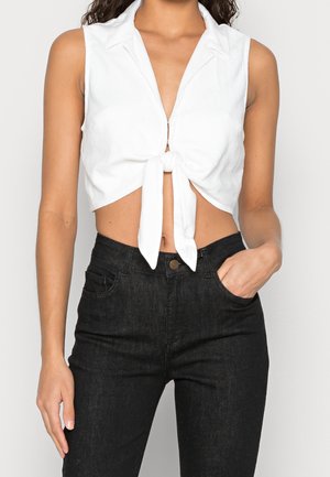 Vrouw draagt een mouwloos wit crop top met strik aan de voorkant en een zwarte, high-waisted jeans, met één hand in de zak tegen een effen achtergrond.