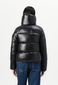 Zwarte pufferjack met een hoge kraag, met een glanzende, gewatteerde textuur en een kort ontwerp. Gecombineerd met blauwe denim jeans.