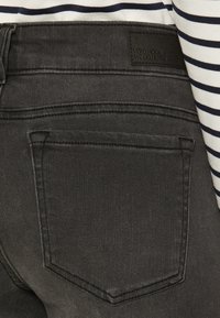 Čierne denimové nohavice s textúrovaným povrchom, obsahujúce malú zadnú kapsu a hnedú nášivku s nápisom "VERO MODA WE DO DENIM." Pásiková košeľa viditeľná nad nimi.