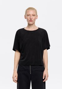Top nero a maniche corte realizzato in morbido tessuto con scollatura rotonda, vestibilità rilassata e texture sottile, abbinato a pantaloni scuri con dettagli zip.