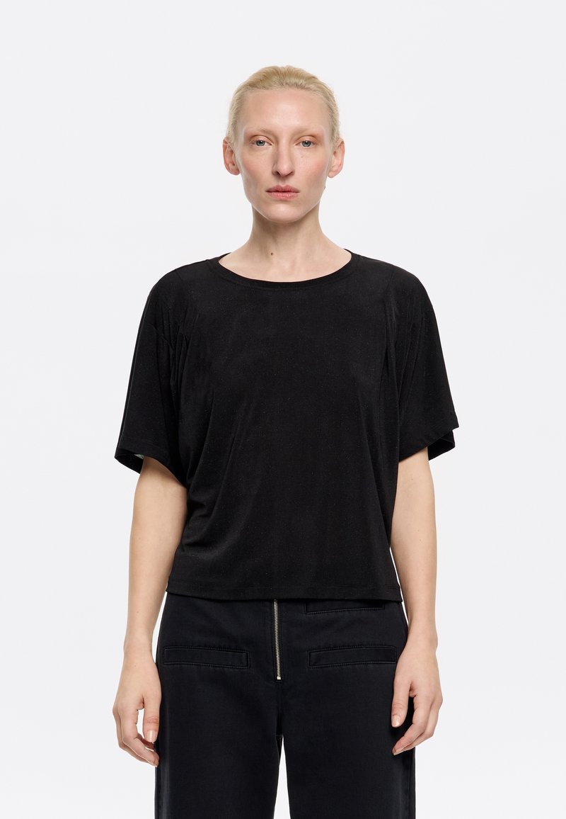 Top nero a maniche corte realizzato in morbido tessuto con scollatura rotonda, vestibilità rilassata e texture sottile, abbinato a pantaloni scuri con dettagli zip.