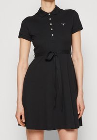 Czarna sukienka polo z bawełny, z kołnierzykiem, krótkimi rękawami i paskiem w talii. Mały detal logo po lewej stronie klatki piersiowej.