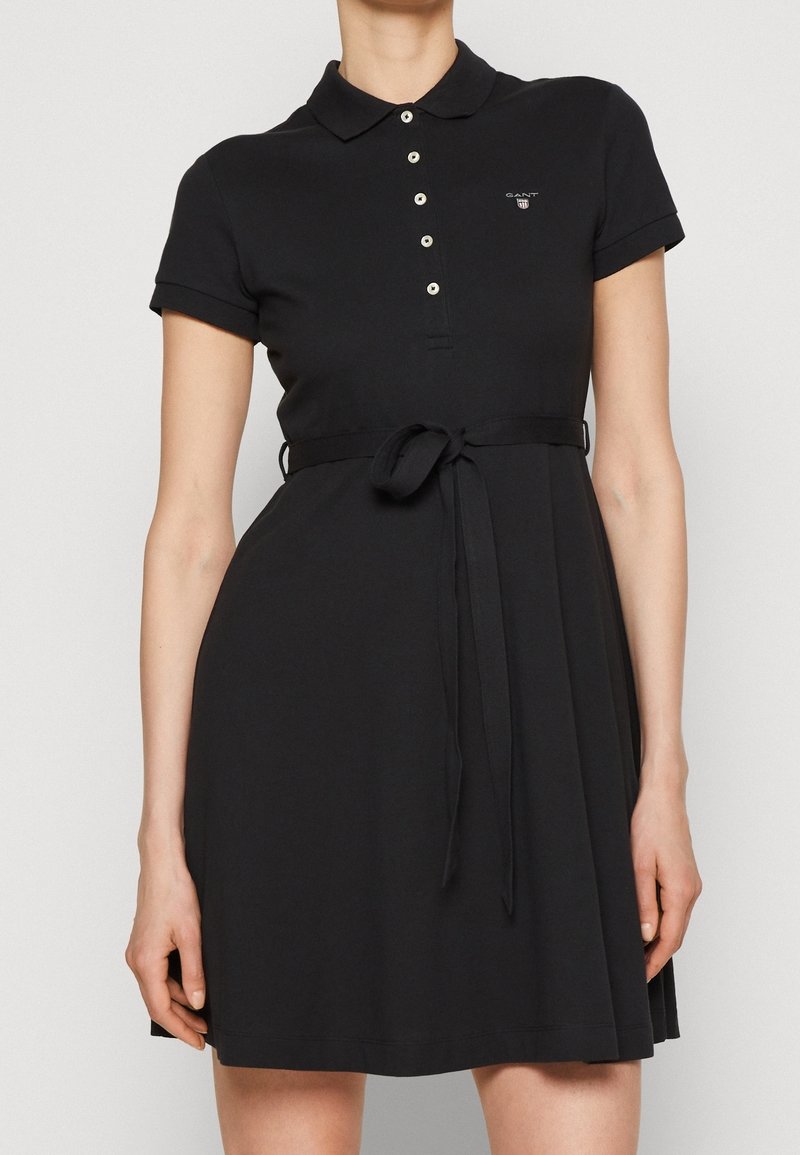 Czarna sukienka polo z bawełny, z kołnierzykiem, krótkimi rękawami i paskiem w talii. Mały detal logo po lewej stronie klatki piersiowej.