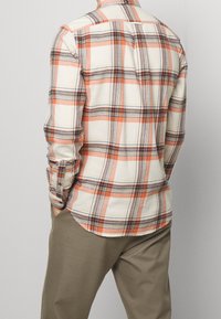 Langarm geruit overhemd in crème, oranje en blauw, met een button-down kraag en ronde zoom. Draagt men met lichtkhaki broeken.