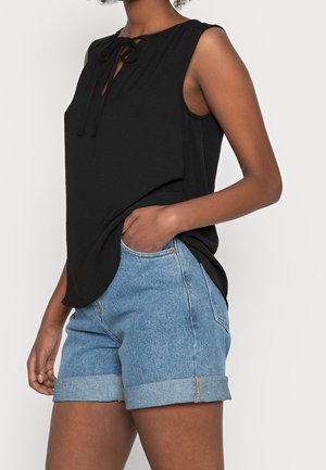 Kvinde iført sort, ærmeløs top med bindesløjfe ved halsen og lyseblå denimshorts med opkrogede kanter, hånd i lommen, foran ensfarvet baggrund.