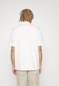 Camicia bianca a maniche corte in cotone, con colletto classico e vestibilità comoda, abbinata a pantaloni beige, vista da dietro.