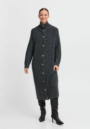 Langärmlige, dunkelgraue Strickjacke mit hohem Kragen, vollen Ärmeln, Frontknöpfen und einem seitlichen Schlitz. Getragen über kniehohen, schwarzen Stiefeln.