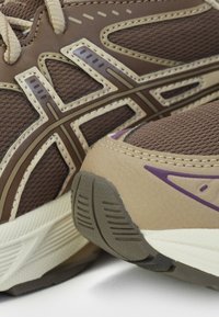 Chaussures de sport marron et beige avec mesh texturé, surpiqûres et semelles à motifs, montrant des détails en gros plan du talon et du côté.