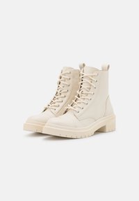 Bottines beiges en cuir avec un bout rond, un laçage à l'avant et un talon épais. Présente des accents de couture et une languette.