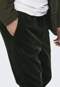 Dark green corduroy pants with vertical ridges, featuring side pockets and a relaxed fit, paired with a white shirt and an olive jacket.