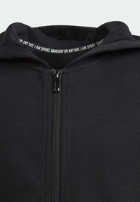 Sort zip-up hoodie med en glat stofstruktur, der har en høj krave og lynlås-lukning. Indvendig label indeholder tekst relateret til sport.