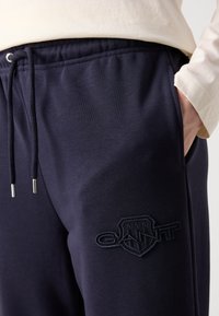 Pantaloni da tuta blu navy realizzati in un tessuto morbido, con vita regolabile tramite cordoncino, tasche laterali e un logo ricamato sulla coscia sinistra.