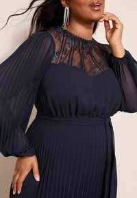 Tumes pleeditud kleit, millel on õhuline pitsist kaelus, pikad pleeditud varrukad ja seotud talje detail. Elegantne tekstuur ja kihiline konstruktsioon.