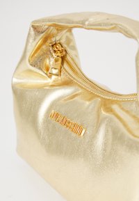 Gullmetallisk håndveske med en slapp design, glidelåslykt og et logo-plater. Har glatt tekstur og et lite hjerteakssent på glidelåsen.