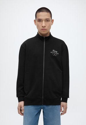 Mężczyzna w czarnej kurtce Tommy Hilfiger na pełny zamek z logo na piersi i jasnoniebieskich dżinsach, stojący na jednolitym białym tle.