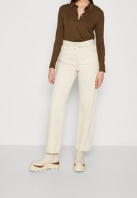 Vaqueros de pierna ancha color beige claro, de talle alto con bolsillos visibles; combinados con una camisa de manga larga marrón oscuro y zapatos de plataforma gruesa.