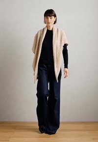 Châle beige drapé sur un haut à manches longues noir, associé à un pantalon en denim foncé évasé. Sol en bois, mur neutre en arrière-plan.