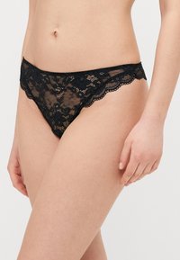 String noir en dentelle avec motif floral, bords festonnés et texture transparente, conçu pour une silhouette ajustée.