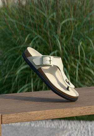 Beige teenslipper met verstelbare metalen gesp, kurken voetbed en zwarte zool, rustend op een houten oppervlak met een wazige groene grasachtergrond.