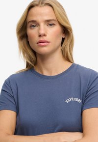 Blå bomuld T-shirt med rund halsudskæring og korte ærmer, med "SUPERDRY" broderet i hvid på venstre brystområde.