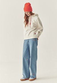 Sudadera con capucha crema y cordones rojos, flores bordadas. Vaqueros de pierna ancha azul claro con detalles florales. Gorro de lana naranja.