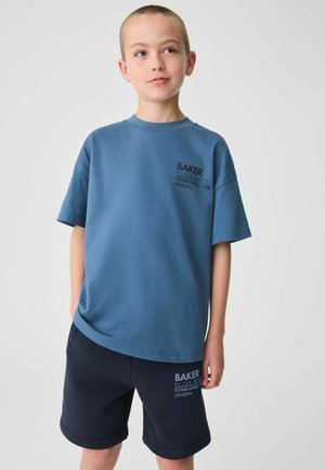 Mladý chlapec s krátkymi vlasmi, oblečený v modrom oversized tričku a tmavých šortkách, oboje s nápisom "Baker by Ted Baker London".