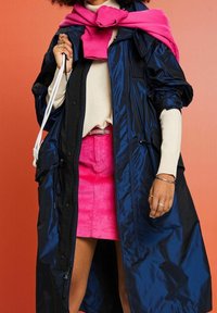 Granatowy, błyszczący długi płaszcz z czarnymi akcentami, w zestawie z jasnobeżowym swetrem i różową spódnicą z corduroy. Dodatki obejmują pierścionki.