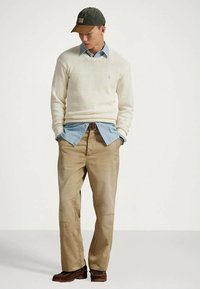 Man med beige byxor, ljusblå skjorta under en krämfärgad tröja, mörkbruna skor och en grön baseballkeps som ser ner med händerna i fickorna.