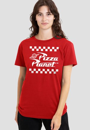 Junge Frau mit langen dunklen Haaren, die ein rotes "Pizza Planet"-T-Shirt und schwarze Hosen trägt, steht vor einem schlichten hellen Hintergrund.
