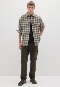 Camisa de franela beige con patrón de cuadros en negro y azul oscuro, usada sobre una camiseta negra. Pantalones de pana verde oliva y zapatillas oscuras.