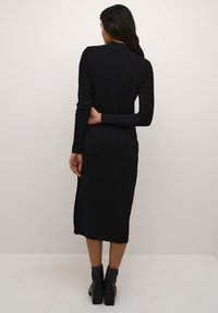 Schwarzes, geripptes Langarmkleid mit hohem Kragen, taillierter Silhouette und seitlichem Schlitz, kombiniert mit klobigen, schwarzen Knöchelstiefeln.