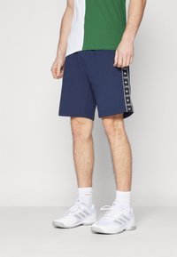 Man wearing navy blue Lacoste shorts with logo stripe, white athletic socks, white Adidas sneakers, and a green and white shirt standing on gray floor.