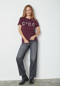 Bordo T-shirt su kurčių „C°CO“ užrašą, derinamas su tamsiai pilkais tiesiais džinsais ir juodais auliniais batais. Lygi medžiaga ir kasualus stilius.