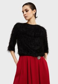 Čierny chlpatý top s trojštvrťovými rukávmi v kombinácii s červenou plisovanou sukňou, ktorá má ozdobnú kvetinovú brošňu na páse.