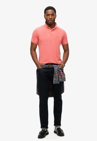 Camisa de polo rosa con cuello, jeans oscuros y mocasines negros. Una camisa de cuadros atada a la cintura añade un detalle en capas, mejorando la textura.