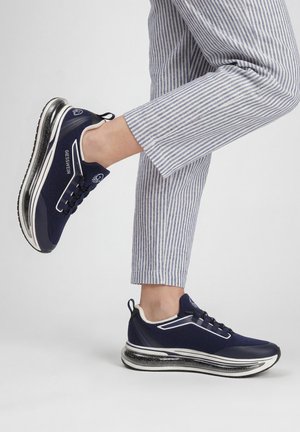 Baskets bleu marine avec semelles épaisses en coussin d’air transparent portées avec un pantalon court rayé bleu clair sur fond clair.