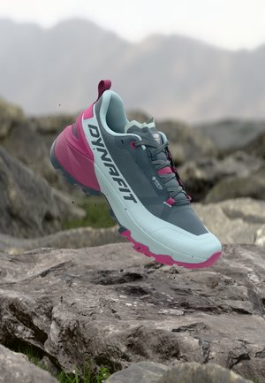 Blue and pink Dynafit trail running shoe floating above rocky terrain with blurred mountain background.
