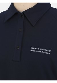 Polo shirt blu navy realizzato in tessuto morbido con chiusura a bottoni. Presenta un colletto classico e testo ricamato bianco sul petto.