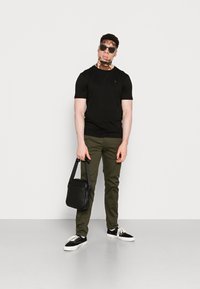 Camiseta negra de manga corta, pantalones verde oliva, bolso cruzado negro y zapatillas en blanco y negro. Gafas de sol en la cara. Fondo claro.