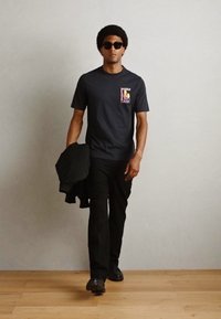 Sort kortærmet t-shirt med grafisk print, parret med sorte bukser og sorte sneakers. Modellen har solbriller på og holder en jakke.