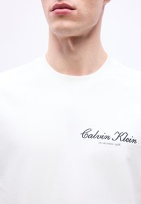 Άνδρας που φοράει λευκό μπλουζάκι με στρογγυλή λαιμόκοψη και κεντημένο λογότυπο «Calvin Klein Established 1968» στο στήθος, το λαιμό και το κάτω μέρος του προσώπου είναι ορατά.