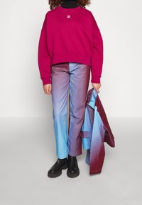 Wino-skarpetkowa bluza z białym logo, jeansy w gradientzie od niebieskiego do fioletowego i czarne botki na platformie. Trzyma czerwoną i niebieską kurtkę.