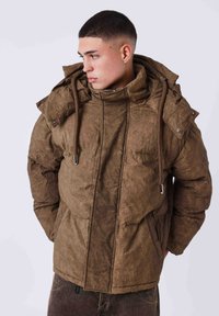 Chaqueta acolchada marrón con cuello alto, capucha y cordones ajustables. Presenta un material texturizado y dos bolsillos frontales con cierres de presión.