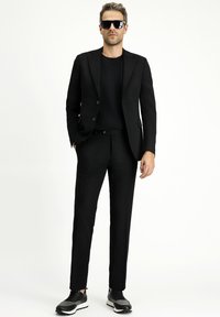 Schwarzer Anzug mit einem taillierten Blazer und Hose, kombiniert mit einem schwarzen T-Shirt und sportlichen schwarz-grauen Sneakers. Übergroße schwarze Sonnenbrille.