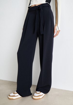 High-waist black wide-leg trousers with fabric belt worn with white and tan sneakers on light floor and white wall background.