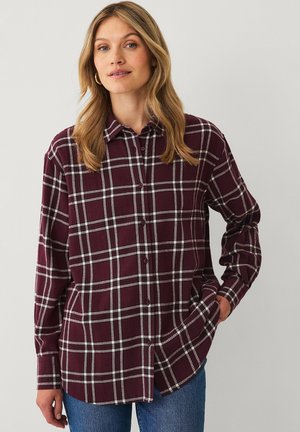 Frau trägt ein burgunder-weiß kariertes Hemd mit langen Ärmeln und blaue Jeans, steht mit einer Hand in der Tasche.