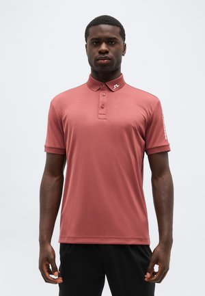 Mladi moški stoji s pogledom naprej, oblečen v prašno rožnato polo majico z gumbano ovratnico in črne hlače na enobarvnem svetlem ozadju.