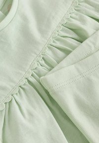 Tissu vert clair avec bord festonné et détail de couture froncée, montrant une partie d'une manche et une couture de corsage.