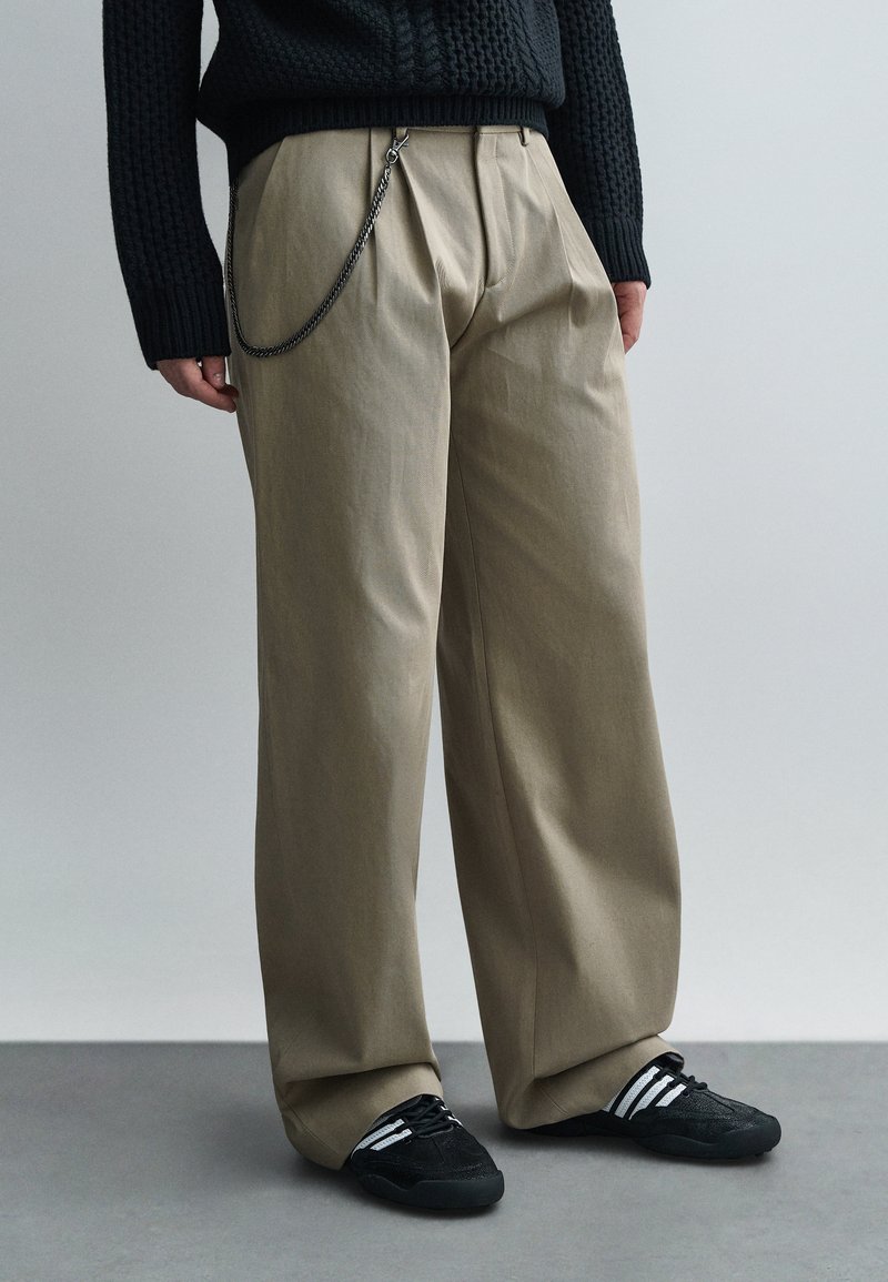 Asmuo dėvi smėlio spalvos platėjančias kelnes su juoda grandinės papuošimu, juodą megztinį ir juodus sportbačius su baltomis juostelėmis, stovintis ant pilkos grindų.